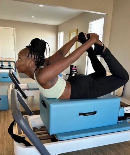 Kayla holding bow pose on the reformer, chest lifted and both hands reaching back to her ankles.